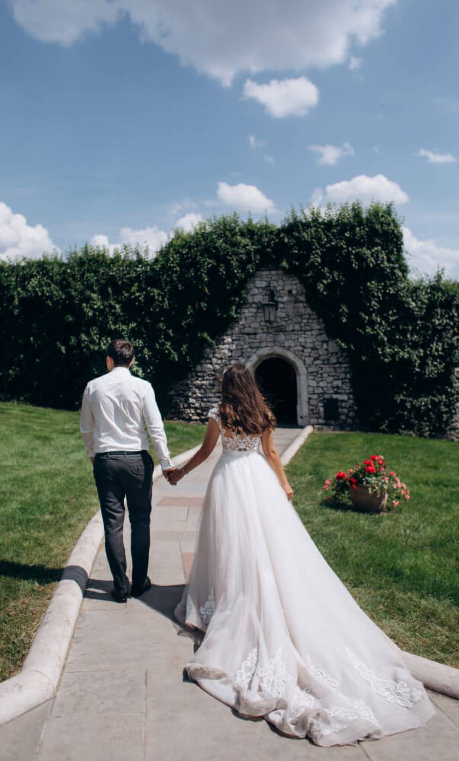 Groom and bride are holding hands and walking to a door in a stone wall at the sunny day